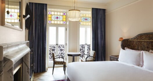 A studio king guest room at the Hotel Chelsea, with a big white bed, a fireplace, and floor-to-ceiling windows with stained-glass details overlooking a balcony.