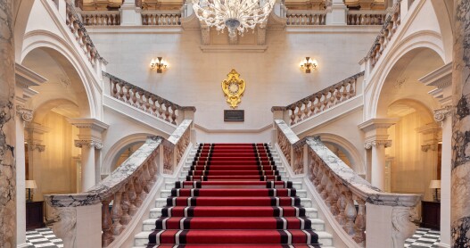 The Grand Staircase at Raffles London at The OWO features stone railings and a red carpet, and is illuminated by a large chandelier.