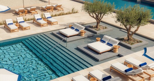 An overhead shot of a bright blue pool lined with white-cushioned wooden lounge chairs and olive trees 